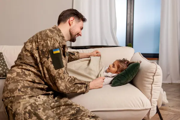happy soldier of Ukrainian army in camouflage uniform returned home and puts his daughter to bed, military dad protects and takes care of child at home, concept of mobilization in Ukraine