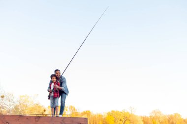 Afrika kökenli Amerikalı bir adam ve çocuk nehirde olta tutan ahşap bir iskelede oturuyor, baba oğluna gölde balık tutmayı öğretiyor, aile hafta sonları dinleniyor ve dinleniyor.