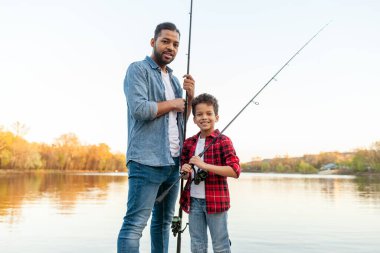 Afrika kökenli Amerikalı bir adam ve çocuk nehirde olta tutan ahşap bir iskelede oturuyor, baba oğluna gölde balık tutmayı öğretiyor, aile hafta sonları dinleniyor ve dinleniyor.