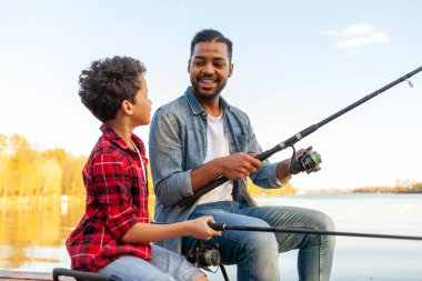 Afrika kökenli Amerikalı bir adam ve çocuk nehirde olta tutan ahşap bir iskelede oturuyor, baba oğluna gölde balık tutmayı öğretiyor, aile hafta sonları dinleniyor ve dinleniyor.