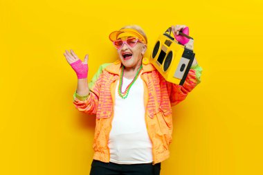 Hippi kıyafetleri içinde komik deli bir büyükanne teypten müzik dinliyor ve sarı izole bir arka planda şarkı söylüyor yaşlı havalı bir kadın partide dans ediyor ve çığlık atıyor.