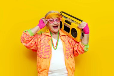 Hippi kıyafetleri içinde komik deli bir büyükanne teypten müzik dinliyor ve sarı izole bir arka planda şarkı söylüyor yaşlı havalı bir kadın partide dans ediyor ve çığlık atıyor.