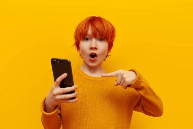 Şok olmuş kızıl saçlı çocuk akıllı telefonu işaret ediyor ve sarı izole edilmiş arka planda şaşırıyor turuncu saç stili ve reklam telefonlu genç hayret içinde.
