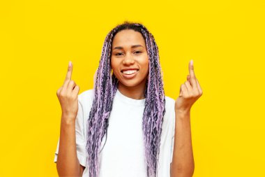 cheerful african american woman with colored dreadlocks showing middle finger on yellow isolated background, hipster girl with purple braids and unique hairstyle showing fuck gesture