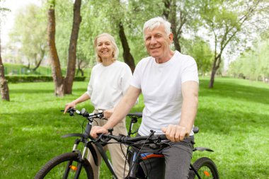 Yaşlı yaşlı çift yazın parkta bisiklete biniyor ve gülümsüyor, yaşlı beyaz saçlı adam ve kadın dışarıda dinleniyor, yaşlı insanlar ormanda bisiklet pratiği yapıyor.