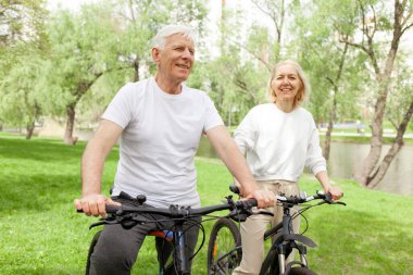 Yaşlı yaşlı çift yazın parkta bisiklete biniyor ve gülümsüyor, yaşlı beyaz saçlı adam ve kadın dışarıda dinleniyor, yaşlı insanlar ormanda bisiklet pratiği yapıyor.