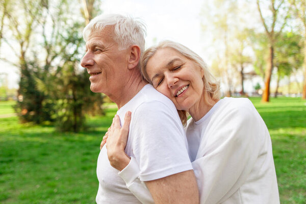 elderly couple of seniors man and woman hugging and smiling in the park outdoors, gray-haired grandparents in white T-shirts in nature
