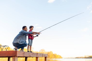 Bir Afrikalı Amerikalı baba ve küçük oğlu bir gölün kıyısındaki ahşap iskelede. Baba, kot pantolon ve mavi gömlek giyer, yere çömelir ve suyu gösterir, oğluna balık tutmayı öğretir. Oğlu elinde oltayla dikkatlice ayakta duruyor, hazır.