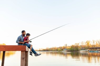 Afrikalı bir Amerikalı baba ve küçük oğlu bir gölün kenarındaki ahşap iskelede oturuyorlar. Kot ceket ve kot pantolon giymiş baba, rıhtımda oturmuş oğlunu tutarken ve oltayı tutmasına yardım ederken, oğlu da kırmızı ekose gömlek ve kot pantolon giyiyordu.