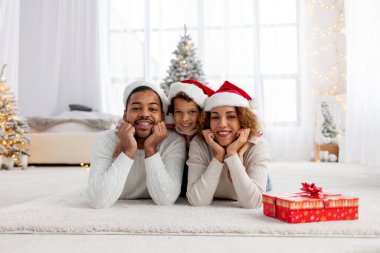 Noel Baba şapkalı Afro-Amerikan ailesi Noel 'de evde sarılıp kameraya bakıyor, genç çocuk anne ve babasıyla yeni yılı kutluyor ve evi dekore ediyor.