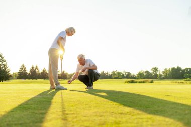 Yaşlı çiftler golf sahasında gün batımında golf oynuyorlar, yaşlı adam ve kadın golf sopalarıyla pratik yapıyorlar, beyaz üniformalı karı koca aktif olarak dinleniyorlar.