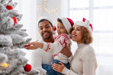 Noel Baba şapkalı Afro-Amerikan ailesi evde birlikte yeni yılı kutluyor ve Noel ağacını süslüyor, genç çocuk anne ve babasıyla yeni yılı kutluyor ve kırmızı oyuncak tutuyor.
