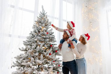 Noel Baba şapkalı Afro-Amerikan ailesi evde birlikte yeni yılı kutluyor ve Noel ağacını süslüyor, genç çocuk anne ve babasıyla yeni yılı kutluyor ve kırmızı oyuncak tutuyor.