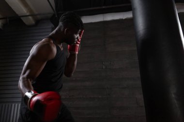 Kırmızı eldiven giyen güçlü Afrikalı Amerikalı boksör spor salonunda kum torbasına vurmaya hazırlanıyor. Boks müsabakasından önce atlet trenleri ve kararlılığını gösteriyor.