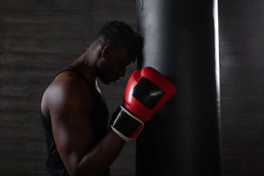 Kırmızı eldiven giyen Afro-Amerikalı bir boksör kafasını kum torbasına yaslamış, düşüncelere dalmış, bir sonraki antrenman için spor salonunun karanlık atmosferinde hazırlanıyor..