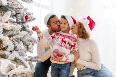Noel Baba şapkalı Afro-Amerikan ailesi evde birlikte yeni yılı kutluyor ve Noel ağacını süslüyor, genç çocuk anne ve babasıyla yeni yılı kutluyor ve kırmızı oyuncak tutuyor.