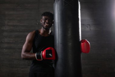 Atletik vücutlu Afrikalı Amerikalı boksör. Kucaklaşma torbası. Kırmızı eldiven takıyor ve spor salonunun karanlık atmosferinde gülümsüyor..