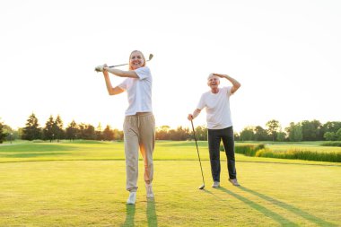 Yaşlı bir çift gün batımında golf oynuyor, yaşlı bir kadın sopayla topa vuruyor ve bir adam ileriye bakıyor, beyaz üniformalı karı koca golf antrenmanı yapıyor.