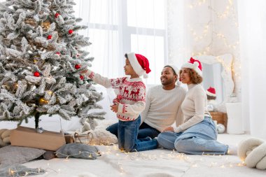 Noel Baba şapkalı Afro-Amerikan ailesi evde birlikte yeni yılı kutluyor ve Noel ağacını süslüyor, genç çocuk anne ve babasıyla yeni yılı kutluyor ve kırmızı oyuncak tutuyor.