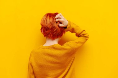 puzzled red-haired teenage boy thinks and chooses on a yellow isolated background, confused child with an orange hairstyle remembers and scratches the back of his head, rear view clipart