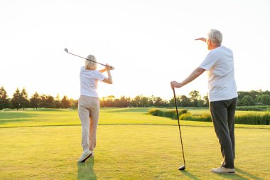 Sahada golf oynayan spor elbiseli yaşlı bir çift. Kadın bir sopa sallıyor, adam arkasından çekim izliyor parlak güneş atmosferi tamamlıyor.. 