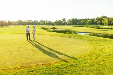 Yaşlı çiftler golf sahasında gün batımında golf oynuyorlar, yaşlı adam ve kadın golf sopalarıyla pratik yapıyorlar, beyaz üniformalı karı koca aktif olarak dinleniyorlar.