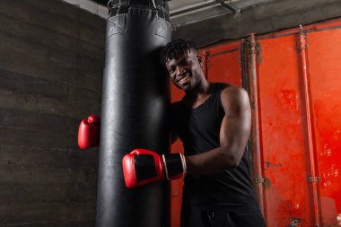Atletik vücutlu Afrikalı Amerikalı boksör. Kucaklaşma torbası. Kırmızı eldiven takıyor ve spor salonunun karanlık atmosferinde gülümsüyor..
