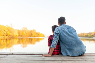 Afrikalı bir Amerikalı baba ve oğlu bir gölün kıyısındaki ahşap iskelede oturuyor, sırtları kameraya dönük. Kot ceket ve kot pantolon giyen baba, kırmızı ekose gömlek giyen oğluna sarılır ve