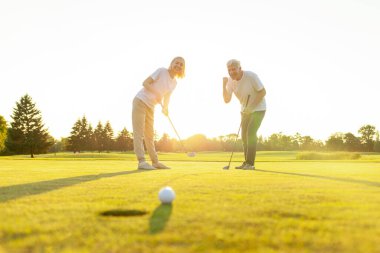 Yaşlı adam ve kadın golfün galibiyetini kutluyor ve el kaldırarak bağırıyor, yaşlı çift gün batımında golf oynuyor ve golf ekipmanlarıyla başarıyı kutluyor.