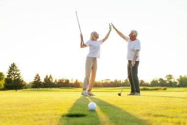 Üniformalı yaşlı çift golf oyununda zaferi ve başarıyı kutluyor ve gün batımında golf oynayan yaşlı adam ve kadına beşlik çakıyor ve açık hava sporları yapıyor.