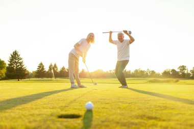 Sahada golf oynayan ve gün batımında zaferi kutlayan yaşlı bir çift yaşlı adam ve kadın golf topunu bir deliğe atıyorlar ve dışarıda birlikte başarıyla eğleniyorlar.
