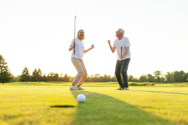 Sahada golf oynayan ve gün batımında zaferi kutlayan yaşlı bir çift yaşlı adam ve kadın golf topunu bir deliğe atıyorlar ve dışarıda birlikte başarıyla eğleniyorlar.