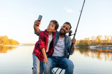 Bir Afrikalı Amerikalı baba ve oğlu gölün kıyısındaki ahşap iskelede oturup balık tutmanın tadını çıkarıyorlar. Baba kot ceket ve kot pantolon giyer, bir elinde olta tutar ve selfie çeker.