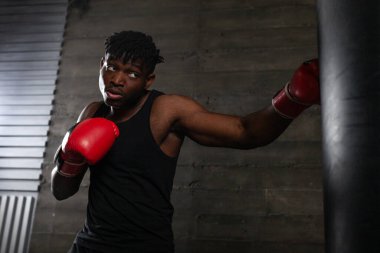 Kırmızı eldiven giyen güçlü Afrikalı Amerikalı boksör spor salonunda kum torbasına vurmaya hazırlanıyor. Boks müsabakasından önce atlet trenleri ve kararlılığını gösteriyor.