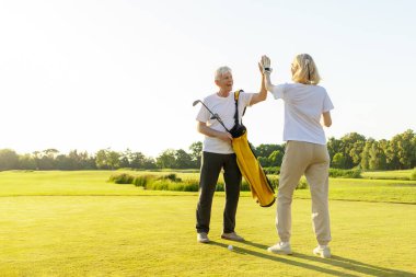 Üniformalı yaşlı çift golf oyununda zaferi ve başarıyı kutluyor ve gün batımında golf oynayan yaşlı adam ve kadına beşlik çakıyor ve açık hava sporları yapıyor.