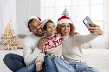 Noel Baba şapkalı Afro-Amerikan aile Noel için evde sarılır ve Noel ağacının yanında selfie çeker. Genç çocuk yeni yılı anne ve babasıyla kutlar ve fotoğraf çeker.