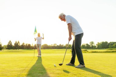 Yaşlı adam ve kadın golf sahasında gün batımında golf oynuyor, yaşlı çift aktif olarak dışarıda eğleniyor ve spor yapıyor, büyükanne ve büyükbaba sopaları tutuyor ve nereye vuracaklarını işaret ediyorlar.