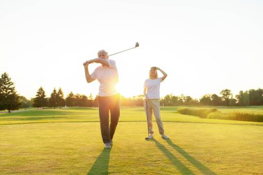 Üniformalı yaşlı çift gün batımında golf oynuyor, yaşlı adam dışarıda golf oynuyor, kadın ileriye bakıyor.