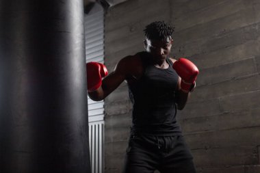 Kırmızı eldiven giyen güçlü Afrikalı Amerikalı boksör spor salonunda kum torbasına vurmaya hazırlanıyor. Boks müsabakasından önce atlet trenleri ve kararlılığını gösteriyor.