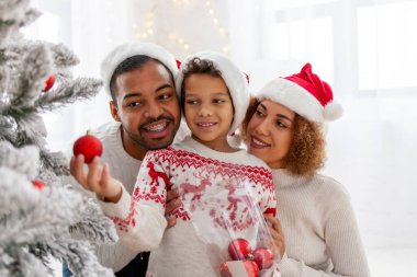 Noel Baba şapkalı Afro-Amerikan ailesi evde birlikte yeni yılı kutluyor ve Noel ağacını süslüyor, genç çocuk anne ve babasıyla yeni yılı kutluyor ve kırmızı oyuncak tutuyor.
