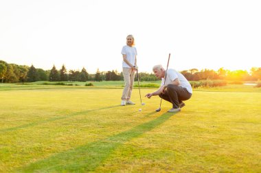 Yaşlı adam ve kadın golf sahasında gün batımında golf oynuyor, yaşlı çift aktif olarak dışarıda eğleniyor ve spor yapıyor, büyükanne ve büyükbaba sopaları tutuyor ve nereye vuracaklarını işaret ediyorlar.