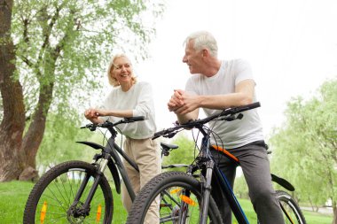 Yaşlı yaşlı çift yazın parkta bisiklete biniyor ve gülümsüyor, yaşlı beyaz saçlı adam ve kadın dışarıda dinleniyor, yaşlı insanlar ormanda bisiklet pratiği yapıyor.