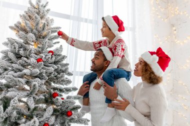 Noel Baba şapkalı Afro-Amerikan ailesi evde birlikte yeni yılı kutluyor ve Noel ağacını süslüyor, genç çocuk anne ve babasıyla yeni yılı kutluyor ve kırmızı oyuncak tutuyor.