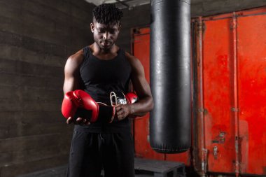 Afrika kökenli Amerikalı bir atlet, konsantrasyonuyla kırmızı bir boks eldiveni çekiyor ve antrenmana hazırlanıyor. Boksör kararlı, kasları gergin, karanlık bir spor salonunda ciddi bir antrenmana hazır..