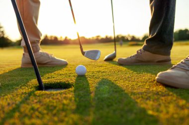 Gün batımında golf oynayan erkek ve kadın, golf topuna ve sopalara yakın çekim, yeşil çimlerin üzerinde insanların ayakları, oyunda yarışıyor ve deliğe vuruyor.