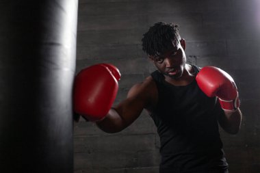 Kırmızı eldiven giyen güçlü Afrikalı Amerikalı boksör spor salonunda kum torbasına vurmaya hazırlanıyor. Boks müsabakasından önce atlet trenleri ve kararlılığını gösteriyor.