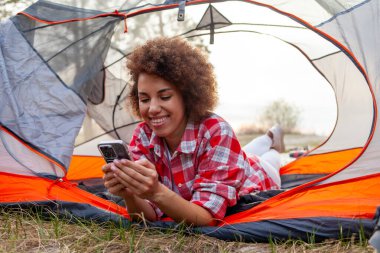 Genç Afrikalı Amerikalı kadın kamp çadırında gölün yakınındaki akıllı telefon kullanarak uzanıyor. Kıvırcık kız turist doğa çadırında cep telefonuyla internette yazı yazıyor.