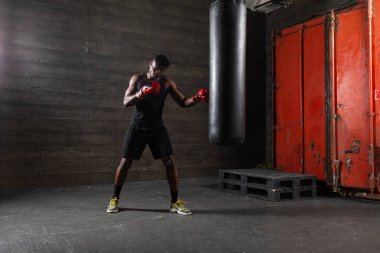 Kırmızı eldiven giyen güçlü Afrikalı Amerikalı boksör spor salonunda kum torbasına vurmaya hazırlanıyor. Boks müsabakasından önce atlet trenleri ve kararlılığını gösteriyor.
