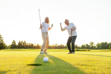 Sahada golf oynayan ve gün batımında zaferi kutlayan yaşlı bir çift yaşlı adam ve kadın golf topunu bir deliğe atıyorlar ve dışarıda birlikte başarıyla eğleniyorlar.