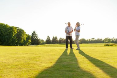 Yaşlı çift golf sahasında golf oynuyor ve gün batımında sohbet ediyor. Yaşlı erkek ve kadın dışarıda dinleniyor ve gülüyor. Emekliler tatilde.